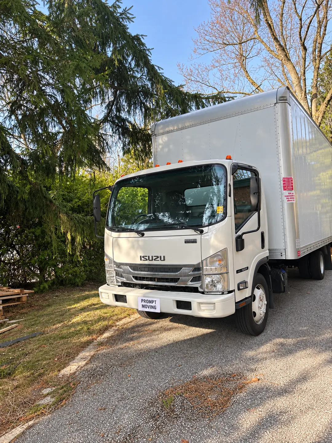Prompt Moving’s white Isuzu truck parked beneath trees, with a branded privacy cover on the plate